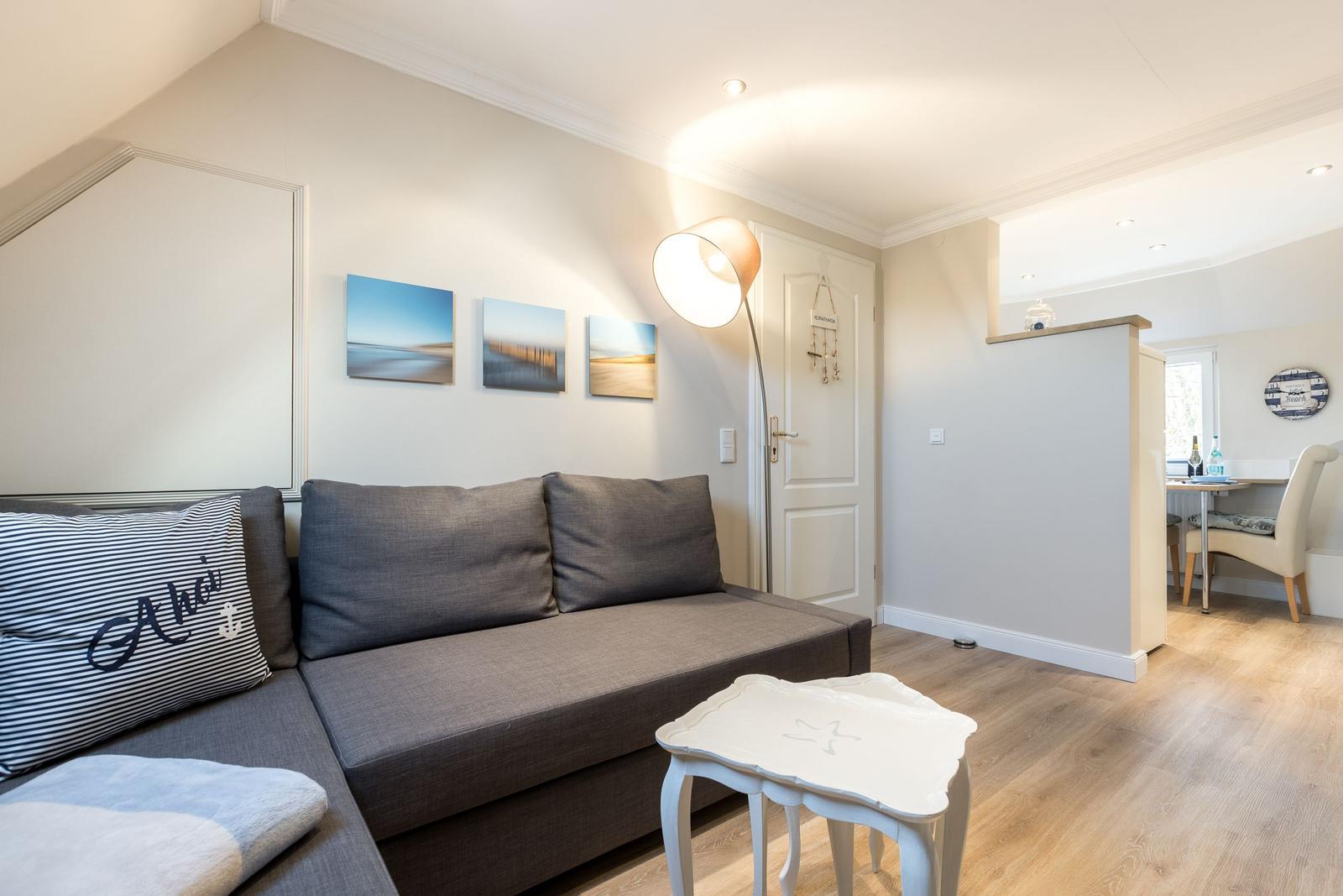 Wohnzimmer mit Sofa, Beistelltisch und Wandbildern. Durchgang zur Küche sichtbar.