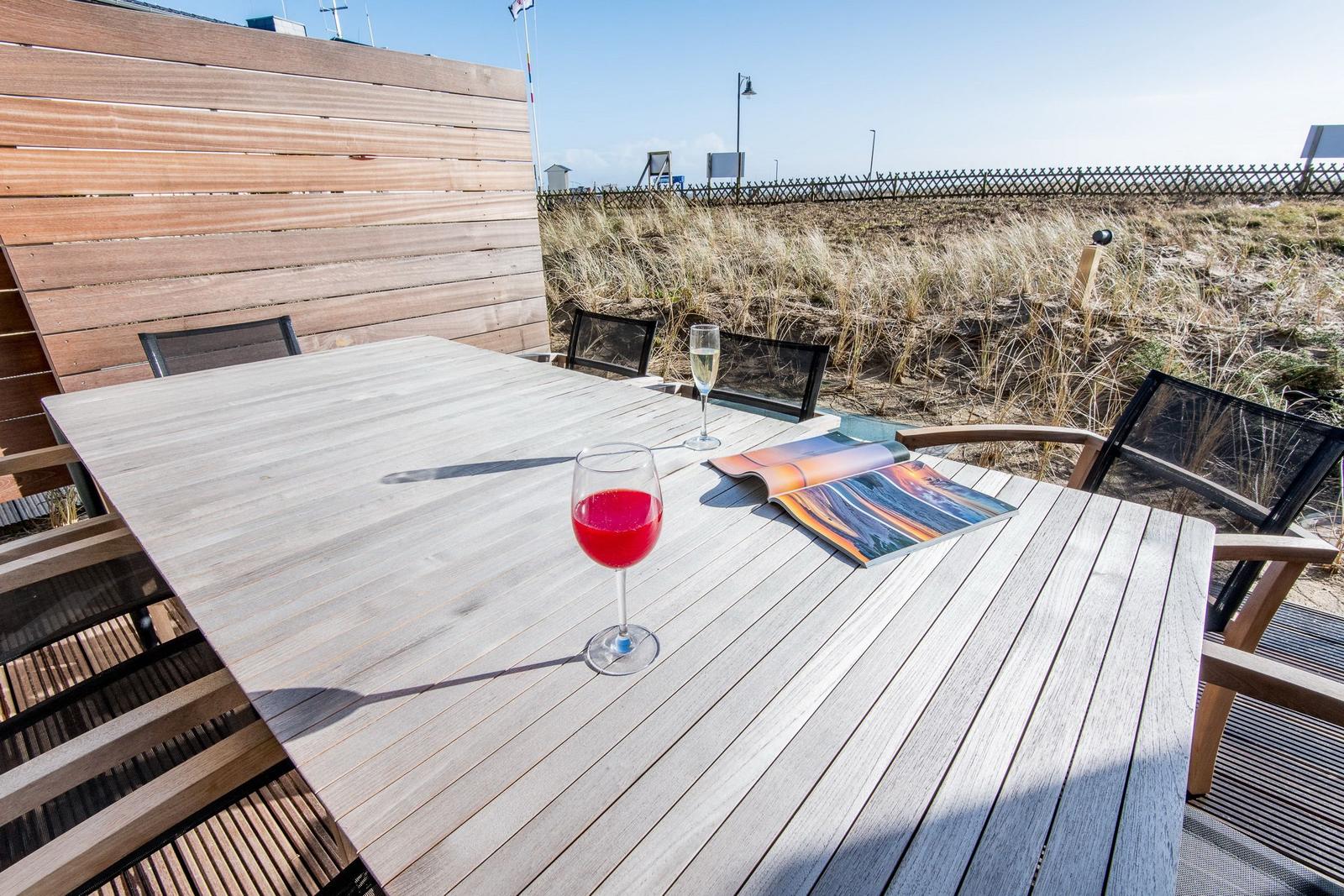 Holztisch mit zwei Gläsern und Zeitschrift auf Terrasse mit Strandhintergrund.