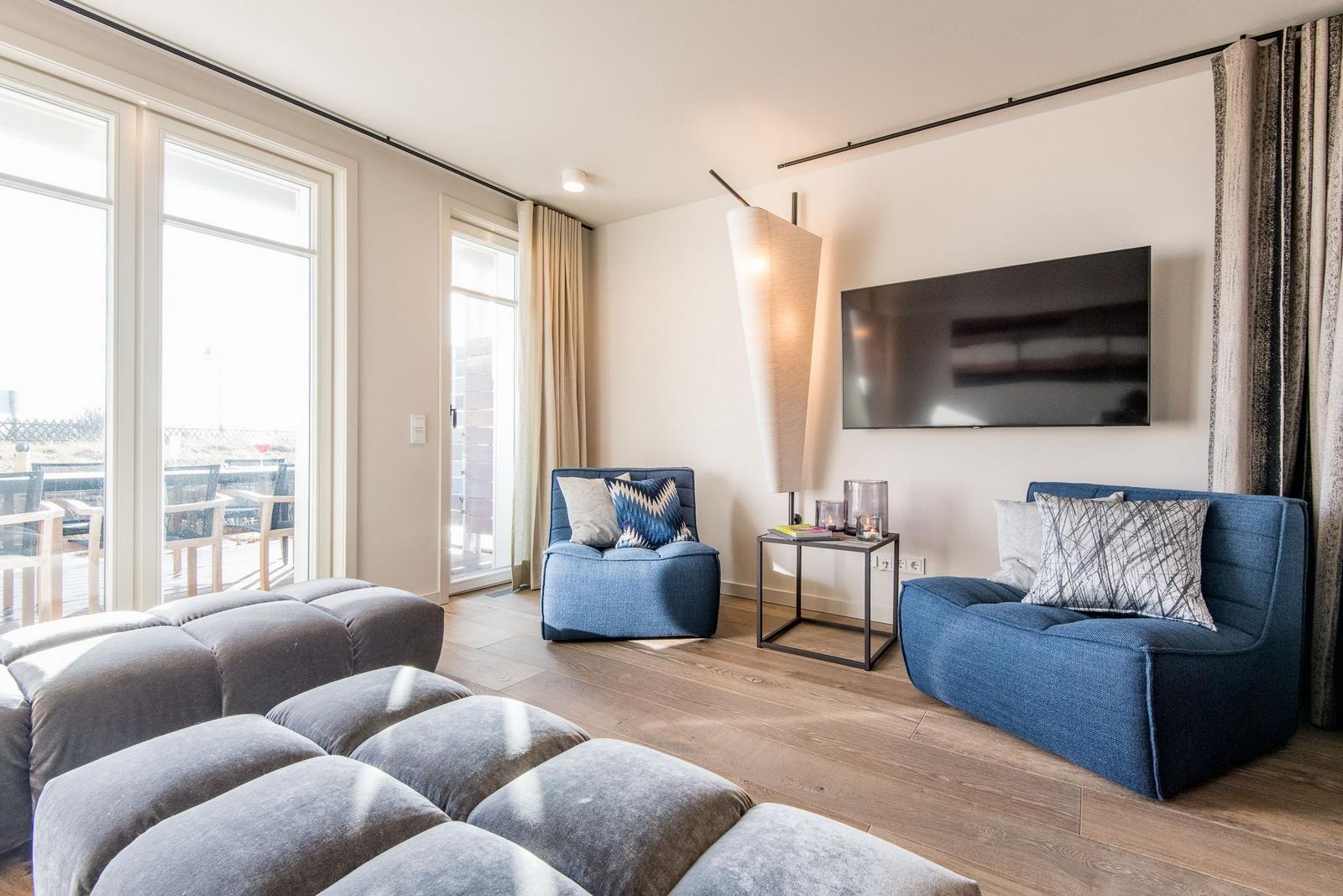 Wohnzimmer mit blauen Sitzmöbeln, Fernseher und großem Fenster zur Terrasse.