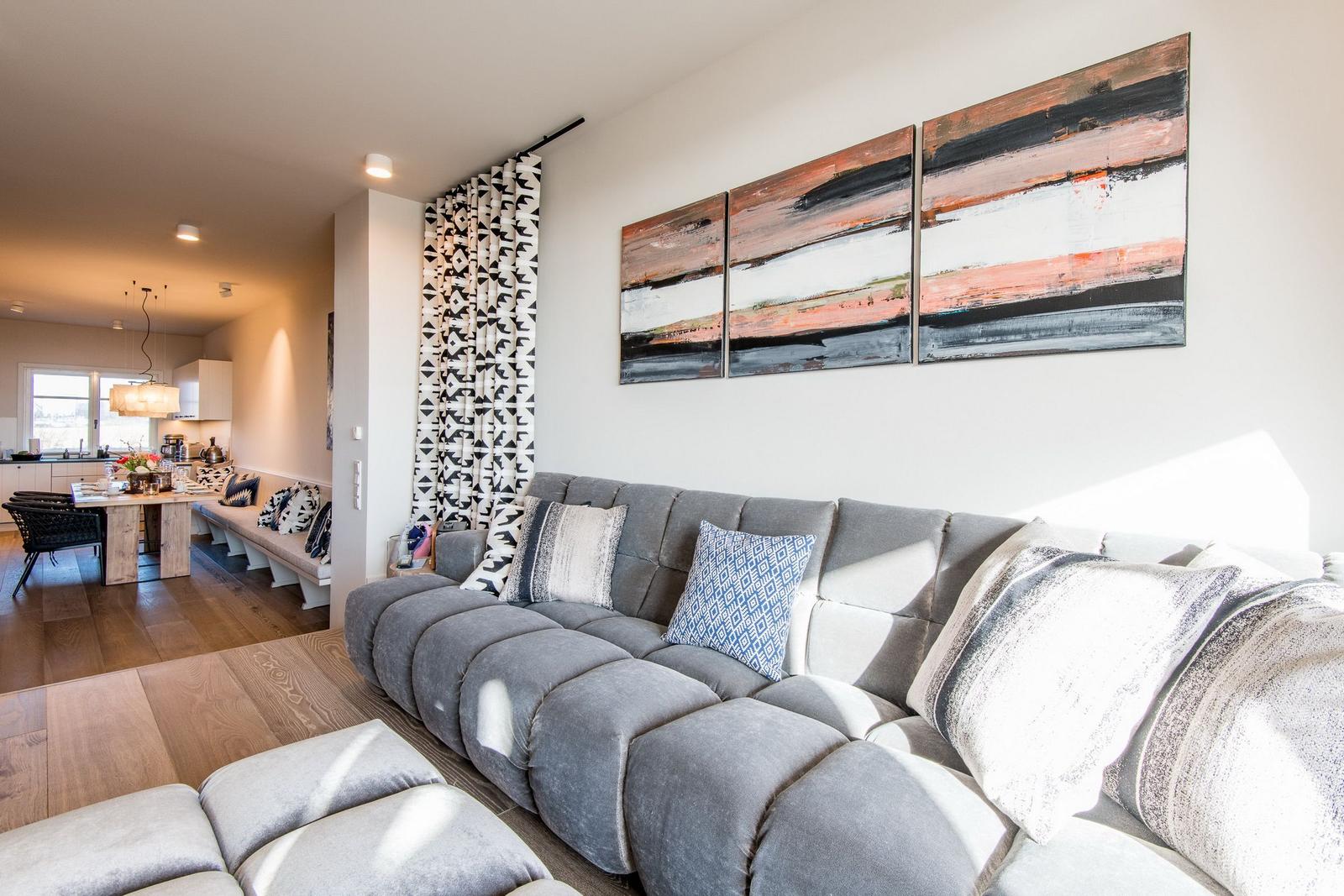 Wohnzimmer mit grauem Sofa, Kissen und Wandbildern. Blick in die Küche mit Esstisch.
