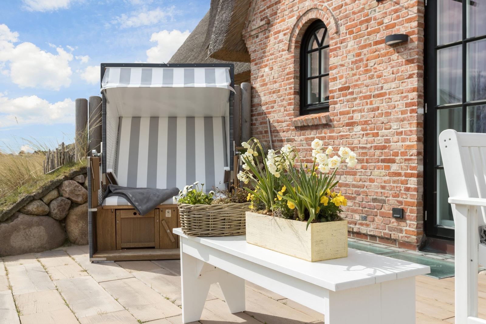 Terrasse mit Strandkorb, Blumen und rotem Backsteinhaus.
