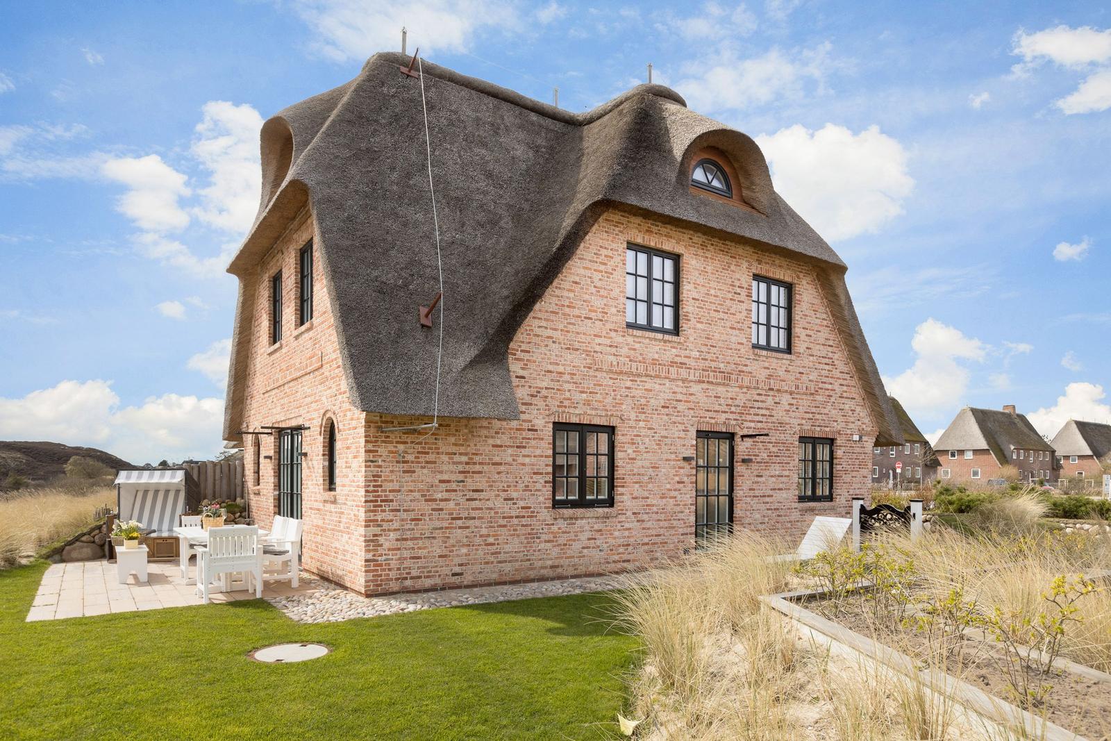 Rotes Backsteinhaus mit Strohdach und Gartenbereich mit weißem Mobiliar.