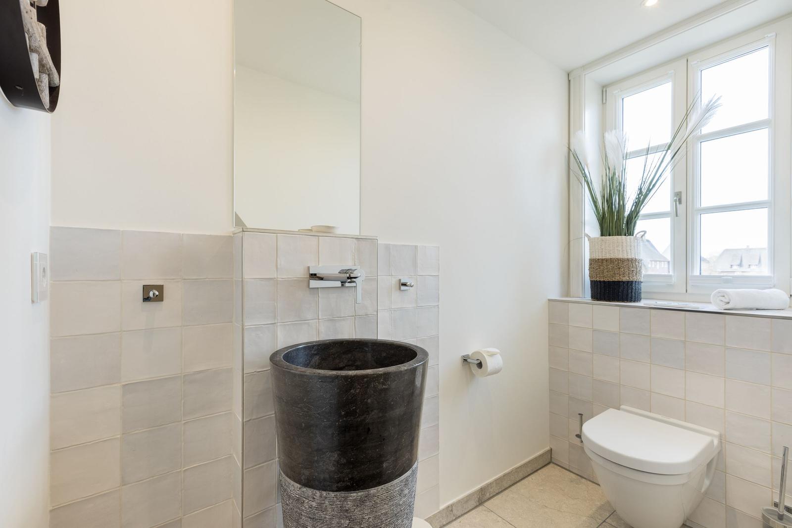 Badezimmer mit Steinwaschbecken, Spiegel und Toilette. Fenster mit Pflanze und Handtuch.