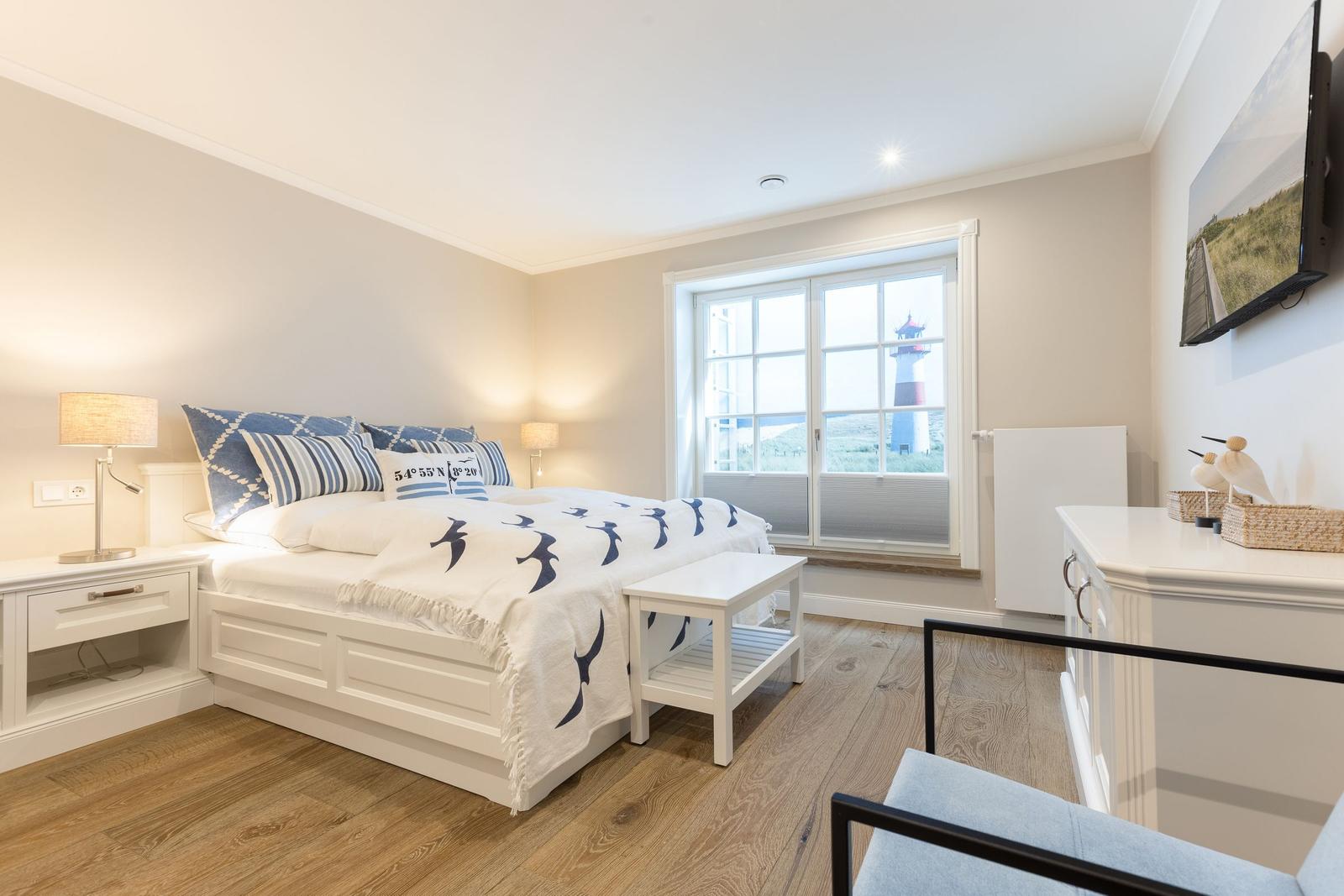 Schlafzimmer mit Blick auf Leuchtturm, weißes Bett, Holzboden und Schreibtisch.