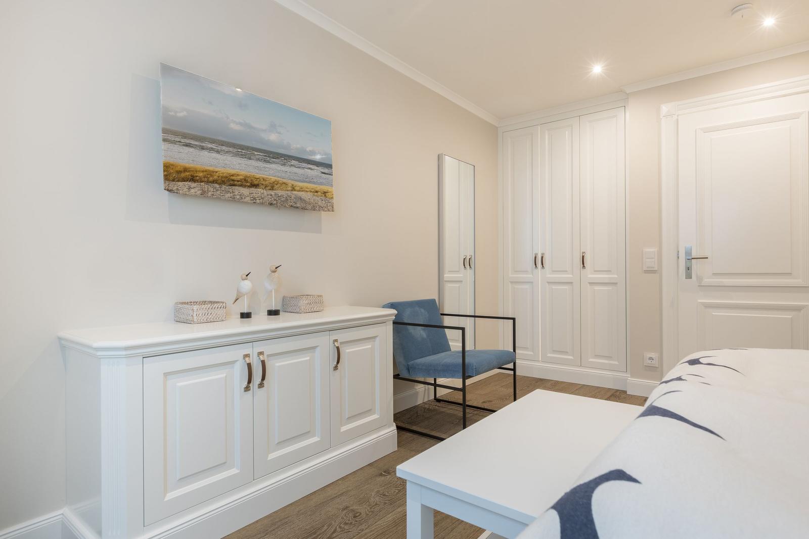 Schlafzimmer mit weißem Schrank, blauer Sitzgelegenheit und Wandbild