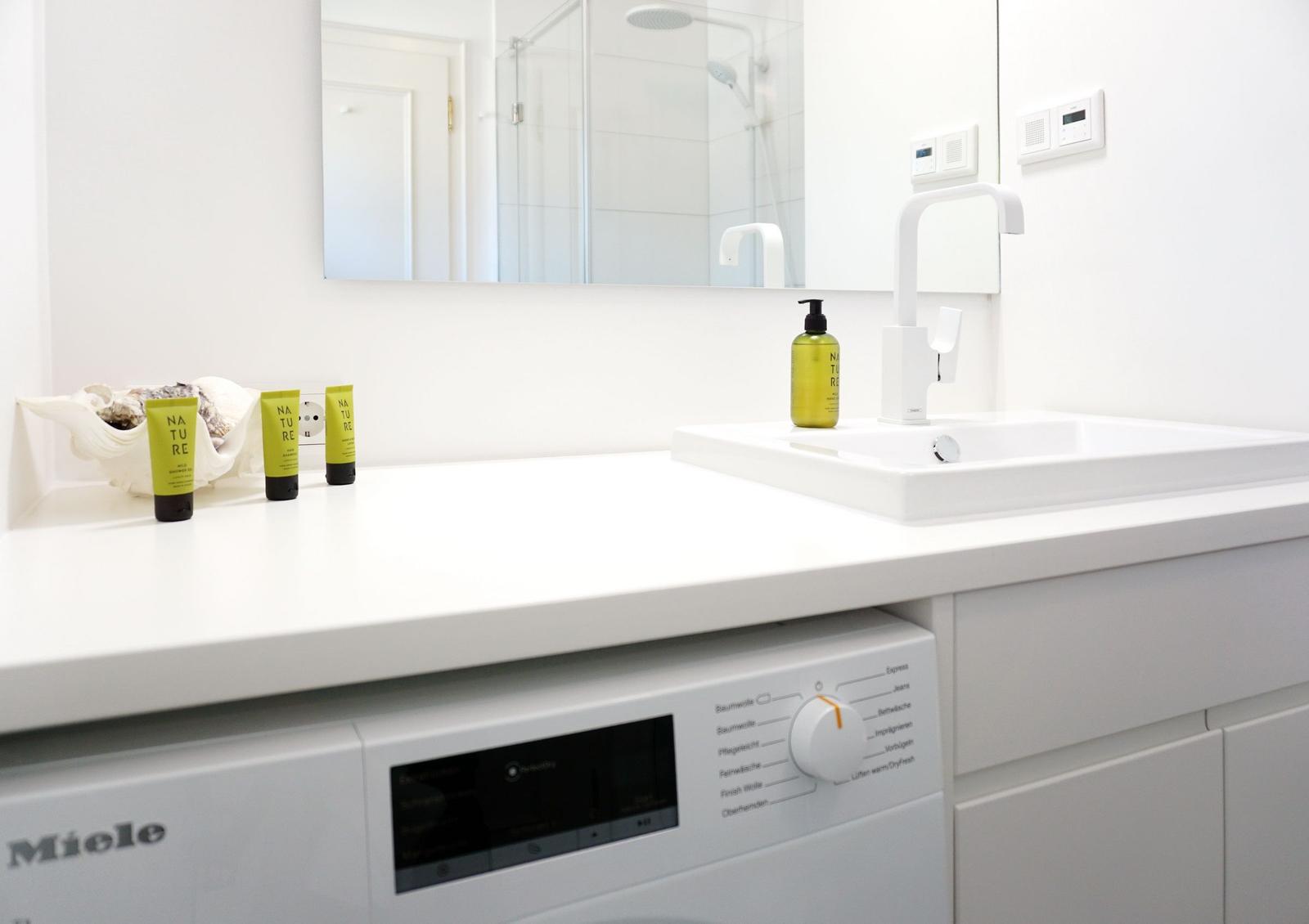 Waschmaschine unter Waschtisch mit Spiegel und Dusche im Hintergrund.