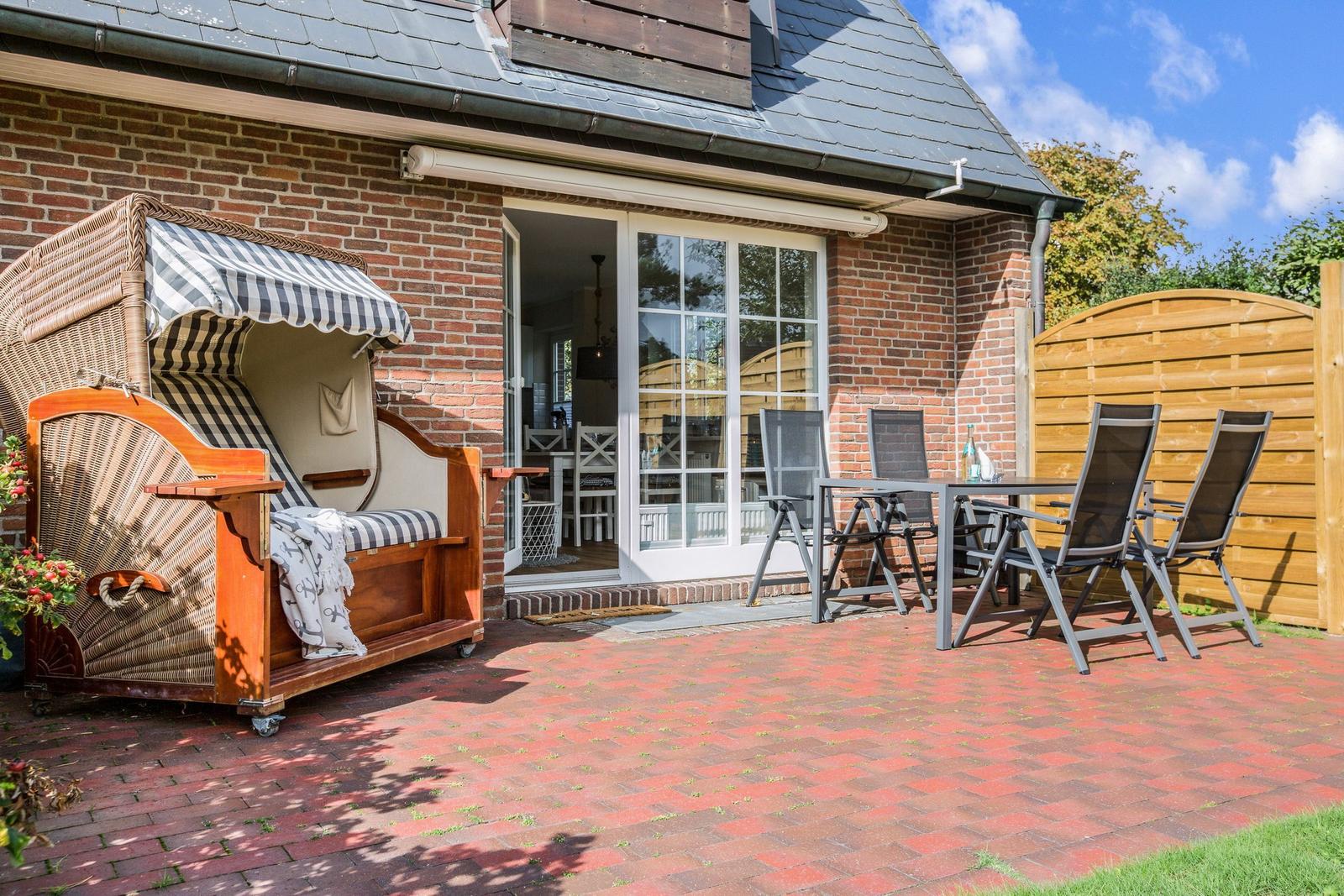 Terrace with sun lounger, table and chairs in front of a house with brick wall.