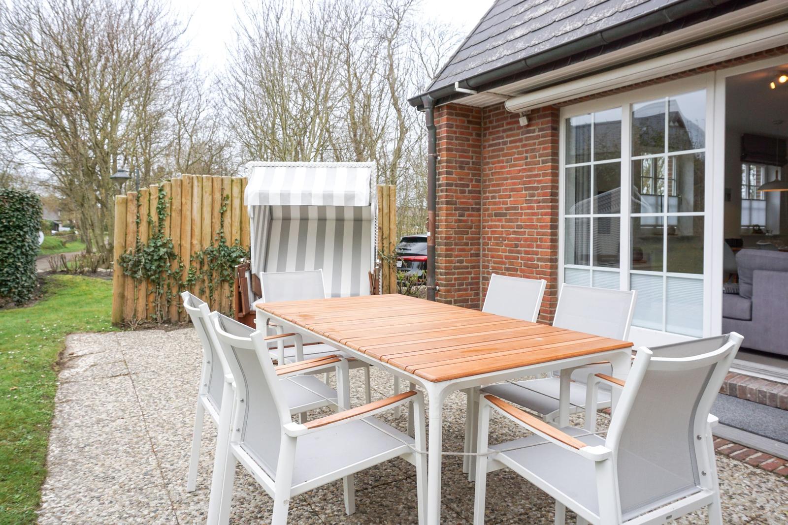 Terrasse mit Tisch, Stühlen und Strandkorb. Hintergrund: Haus mit Glasfront und Holzzaun.