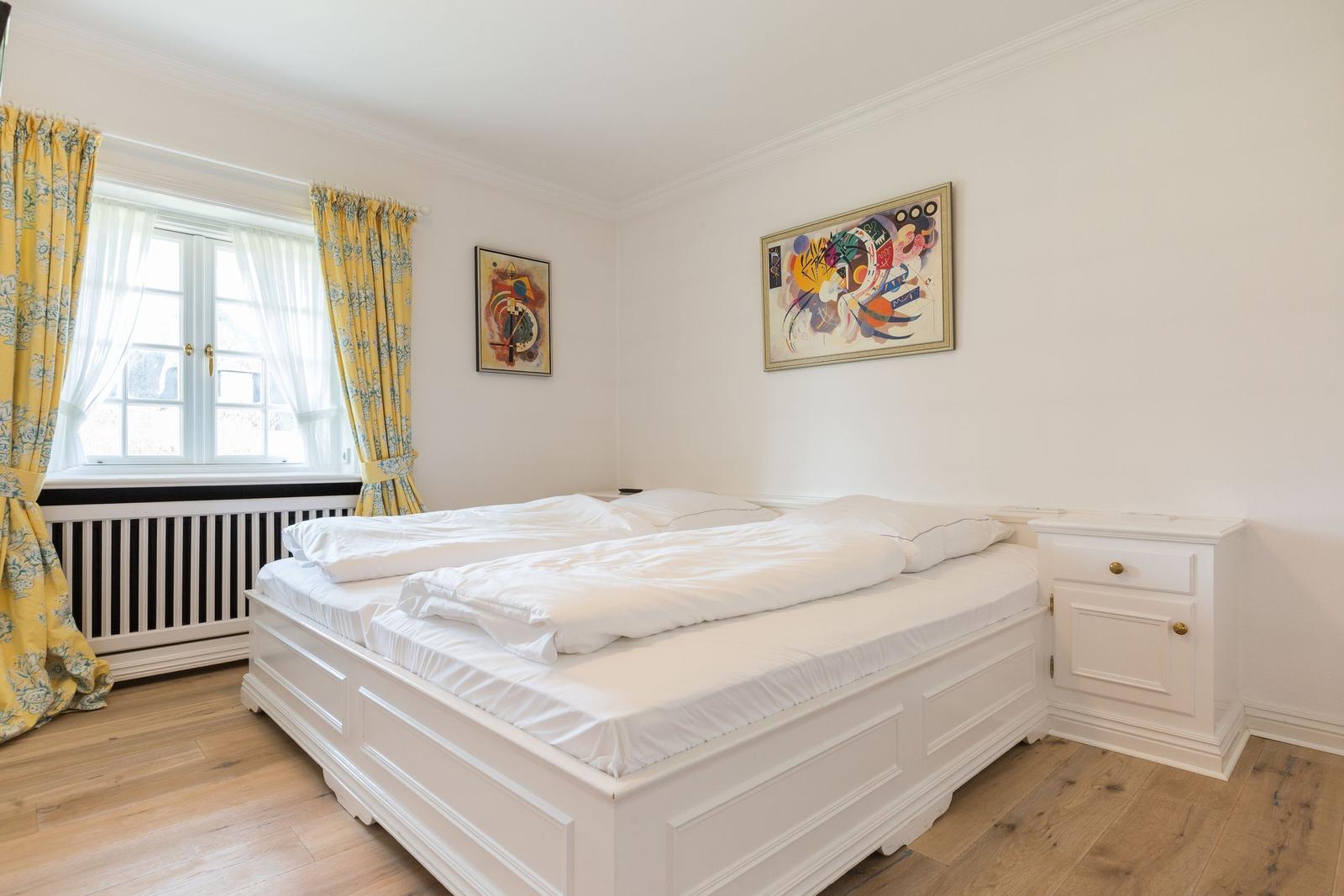 Schlafzimmer mit weißem Bett, gelben Vorhängen und abstraktem Bild an der Wand.