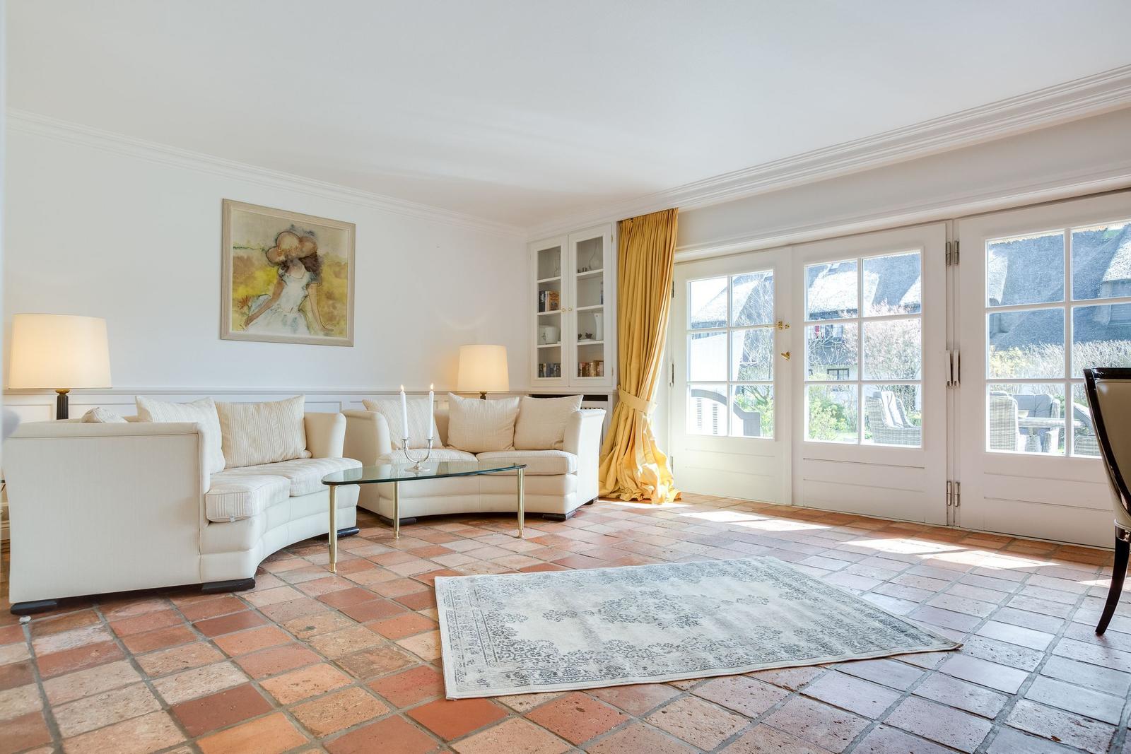 Wohnzimmer mit cremefarbenen Sofas, Glas-Tisch und Terrakottboden. Große Fenster mit gelben Vorhängen.