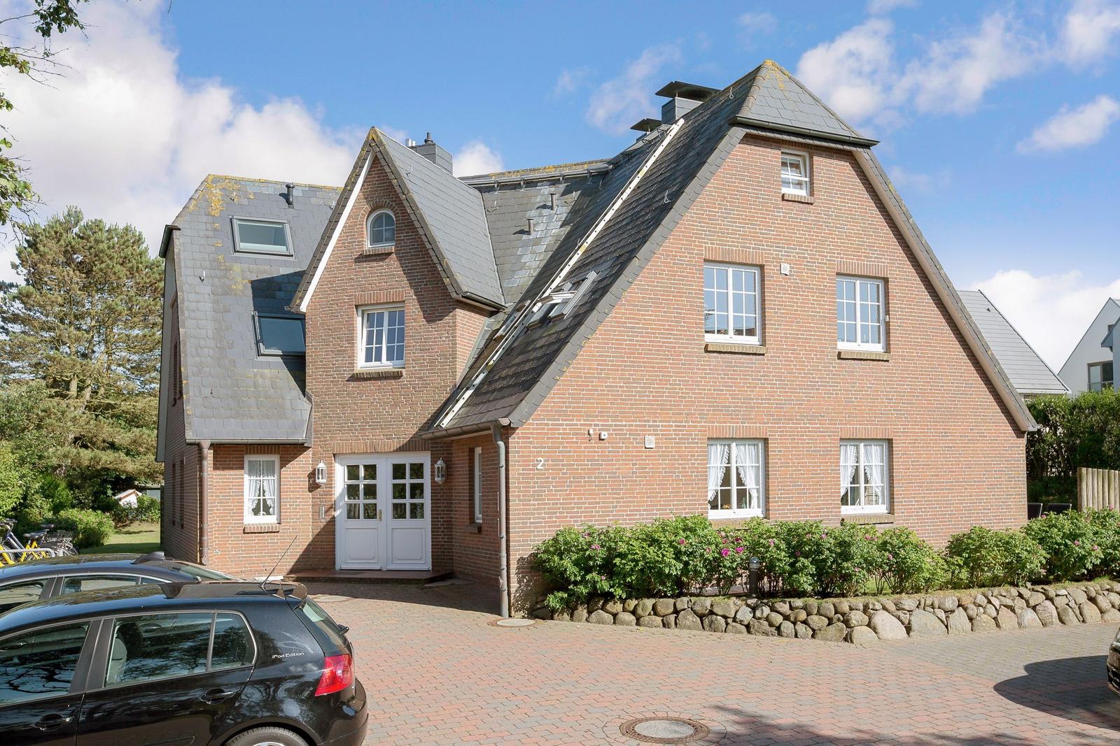 Red brick house with gray roof and white windows. Courtyard with stone wall and paved parking area.