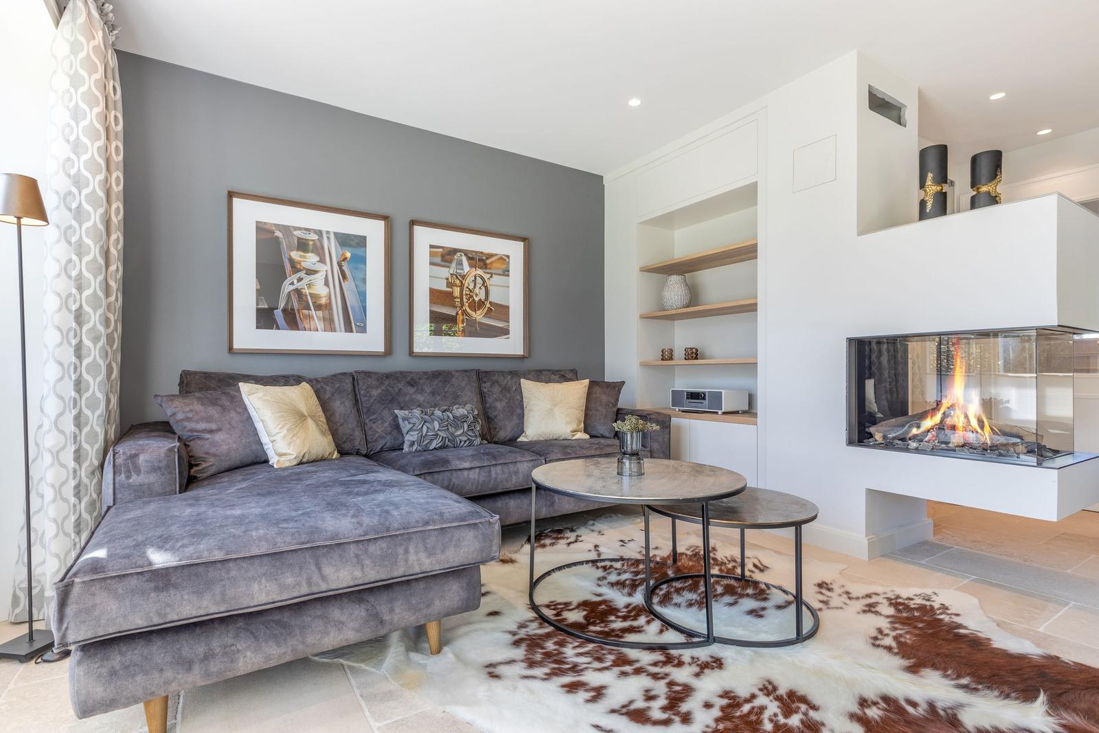 Ein Wohnzimmer mit grauem Sofa, Kamin und runden Tischen auf einem Fellteppich.