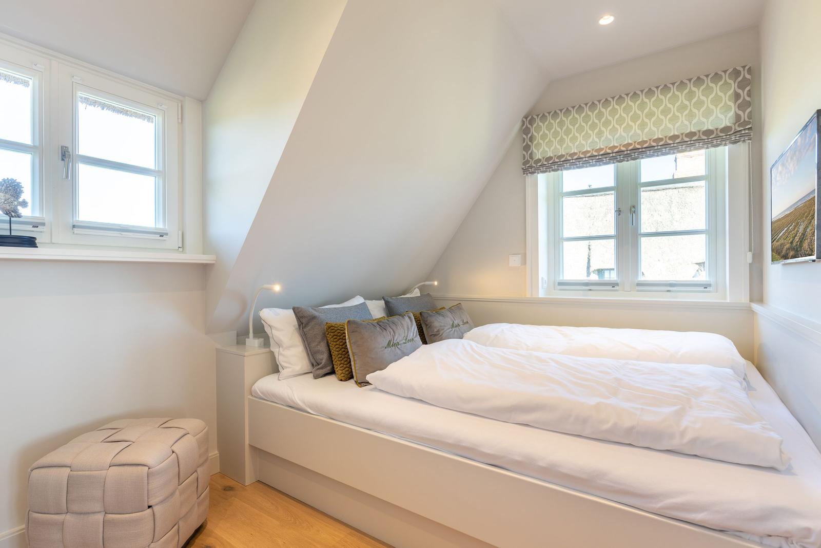 Schlafzimmer mit Dachschrägen, Bett, Fenstern und Sitzhocker.