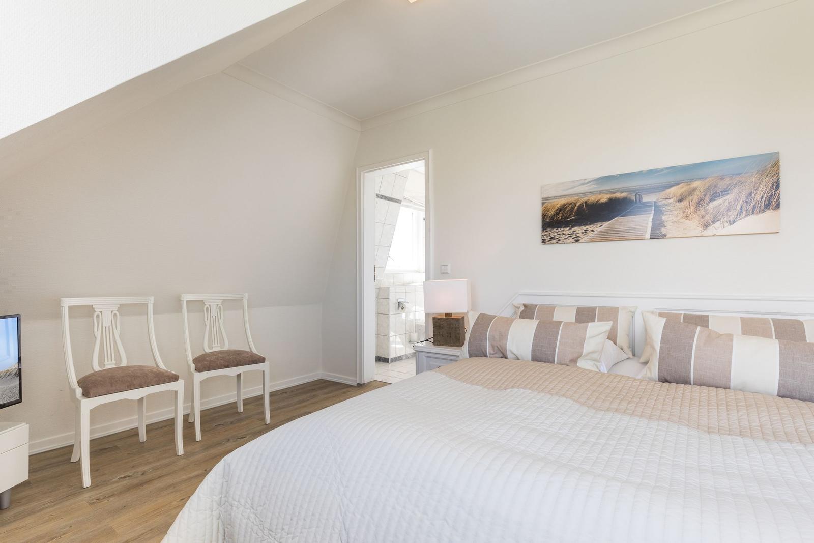 Schlafzimmer mit Bett, zwei Stühlen und Wandbild. Tür führt in Bad.