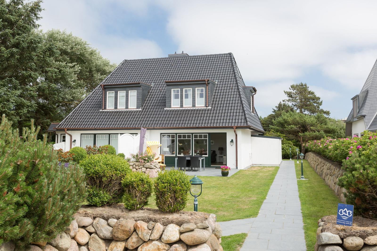 Haus mit dunklem Dach, weißer Fassade und Gartenweg. Terrasse mit Tisch und Stühlen.