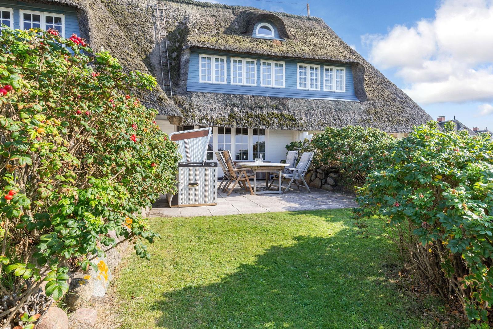 House with thatched roof, terrace with table and chairs, surrounded by grass and shrubs.