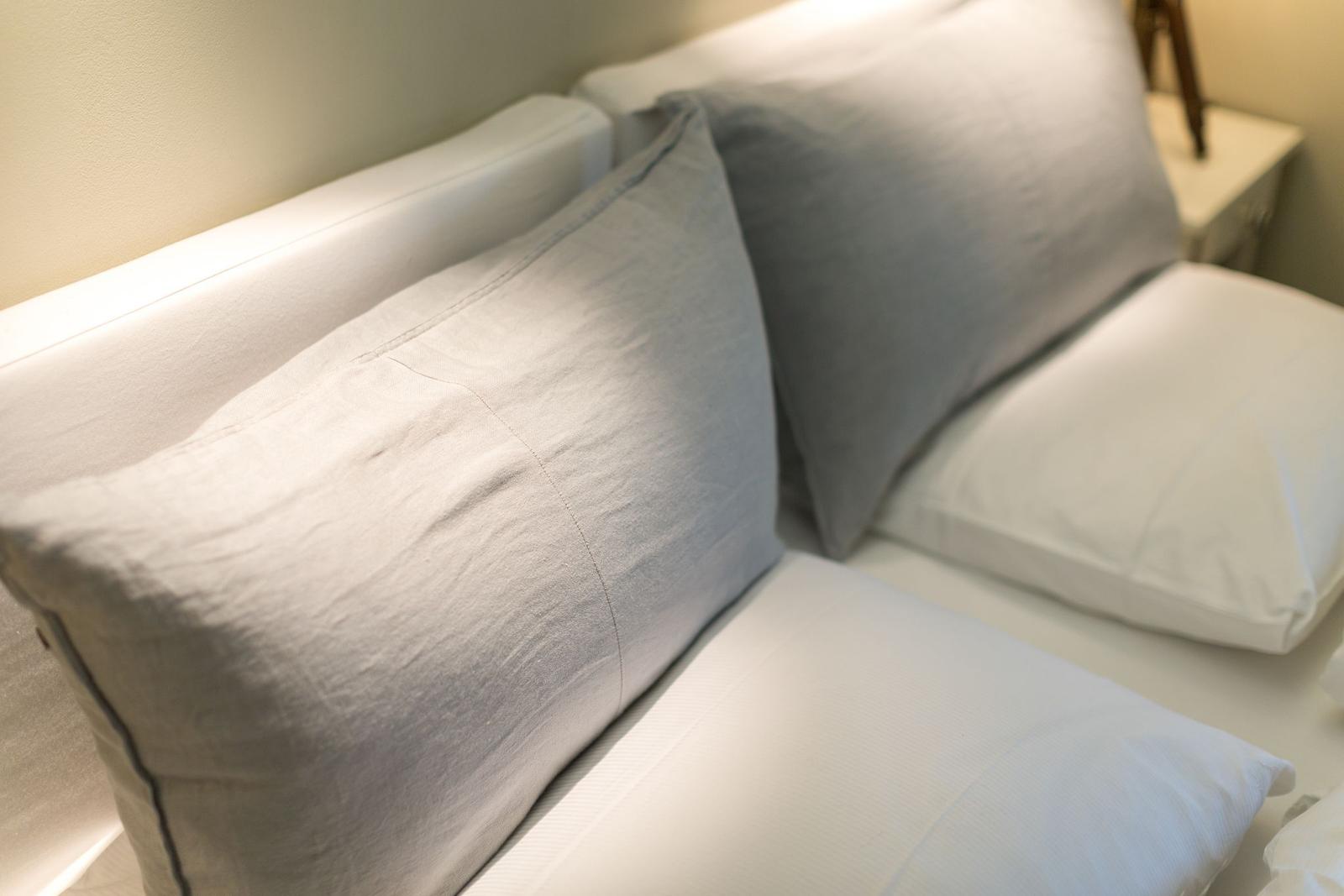 A bed with white sheets and two pillows, one of which is gray.