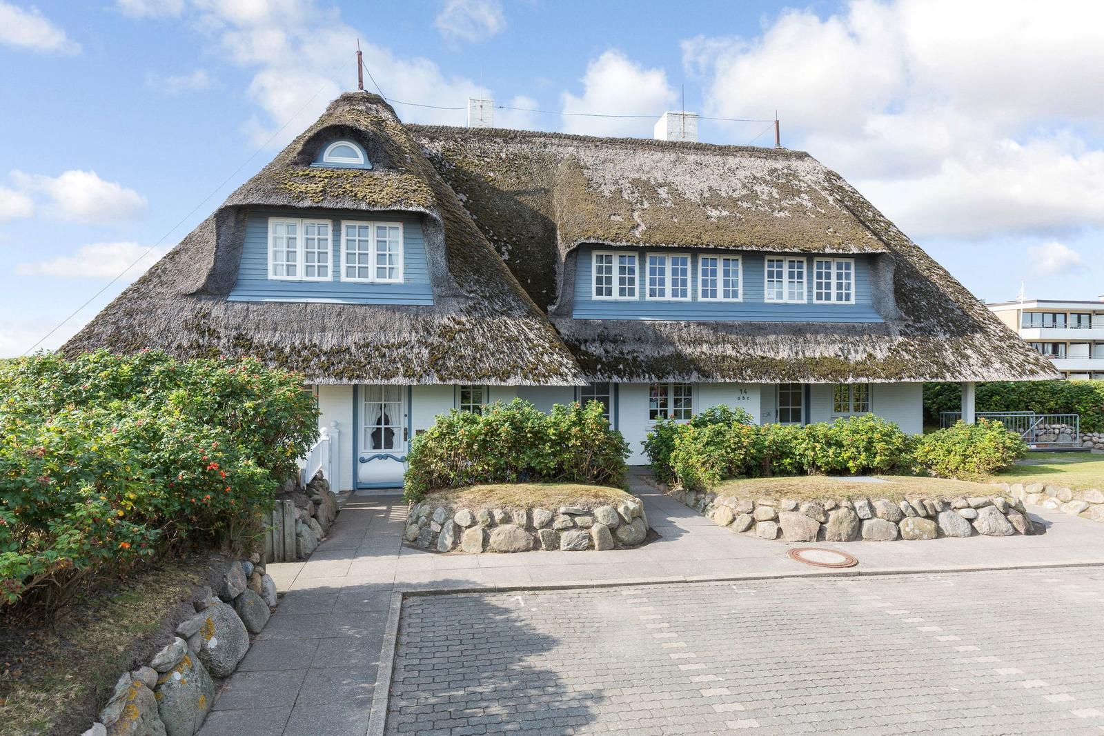 Holiday home with thatched roof and blue windows. Front garden with stone border and shrubs.