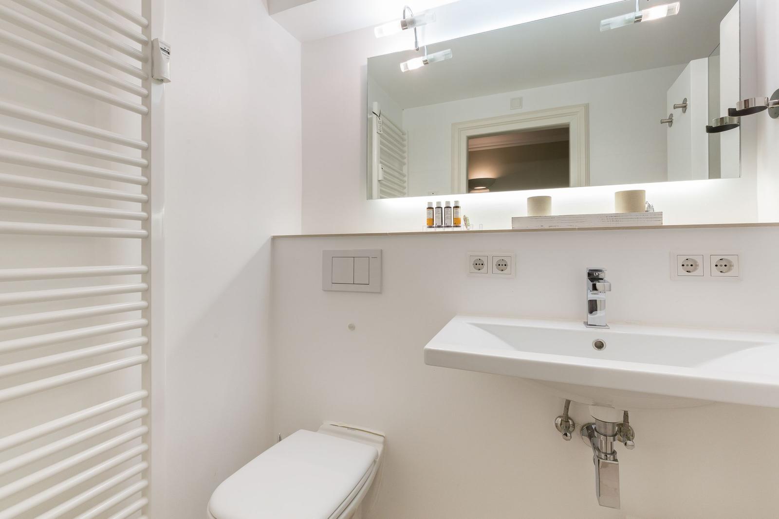 Bathroom with sink, mirror, and toilet. Warm lighting and white walls.
