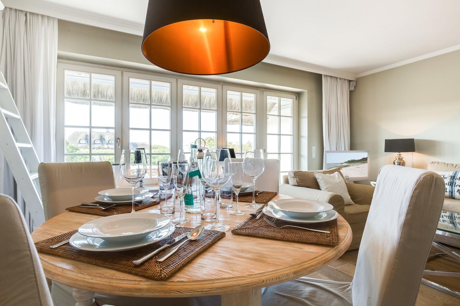 Round table with placemats and glasses. Windows with view. Living area in background.