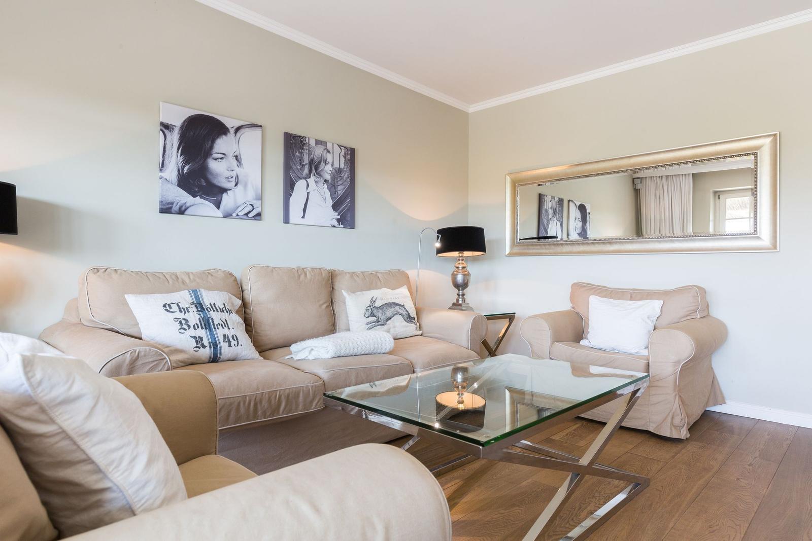 Living room with sofa, armchair, and glass table. Wall art and mirror on the walls.