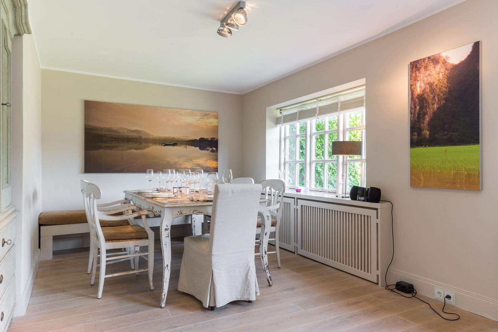 Dining room with table, chairs, and large windows. Wall art shows landscapes.