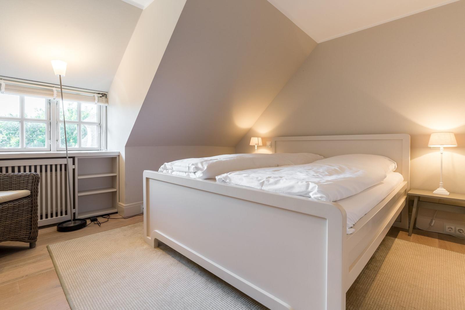 Bedroom with sloped ceiling, white bed, and window with view outside.