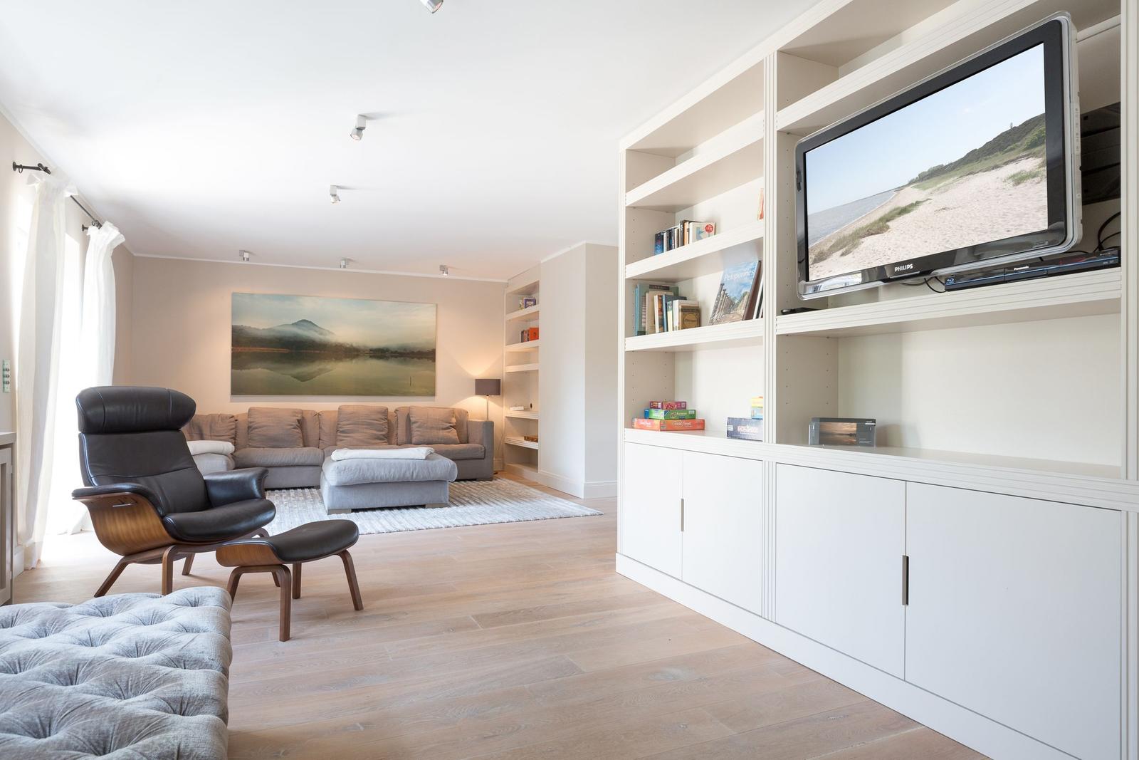 Living room with sofa, chair, and TV area. Wooden floor and bookshelves.