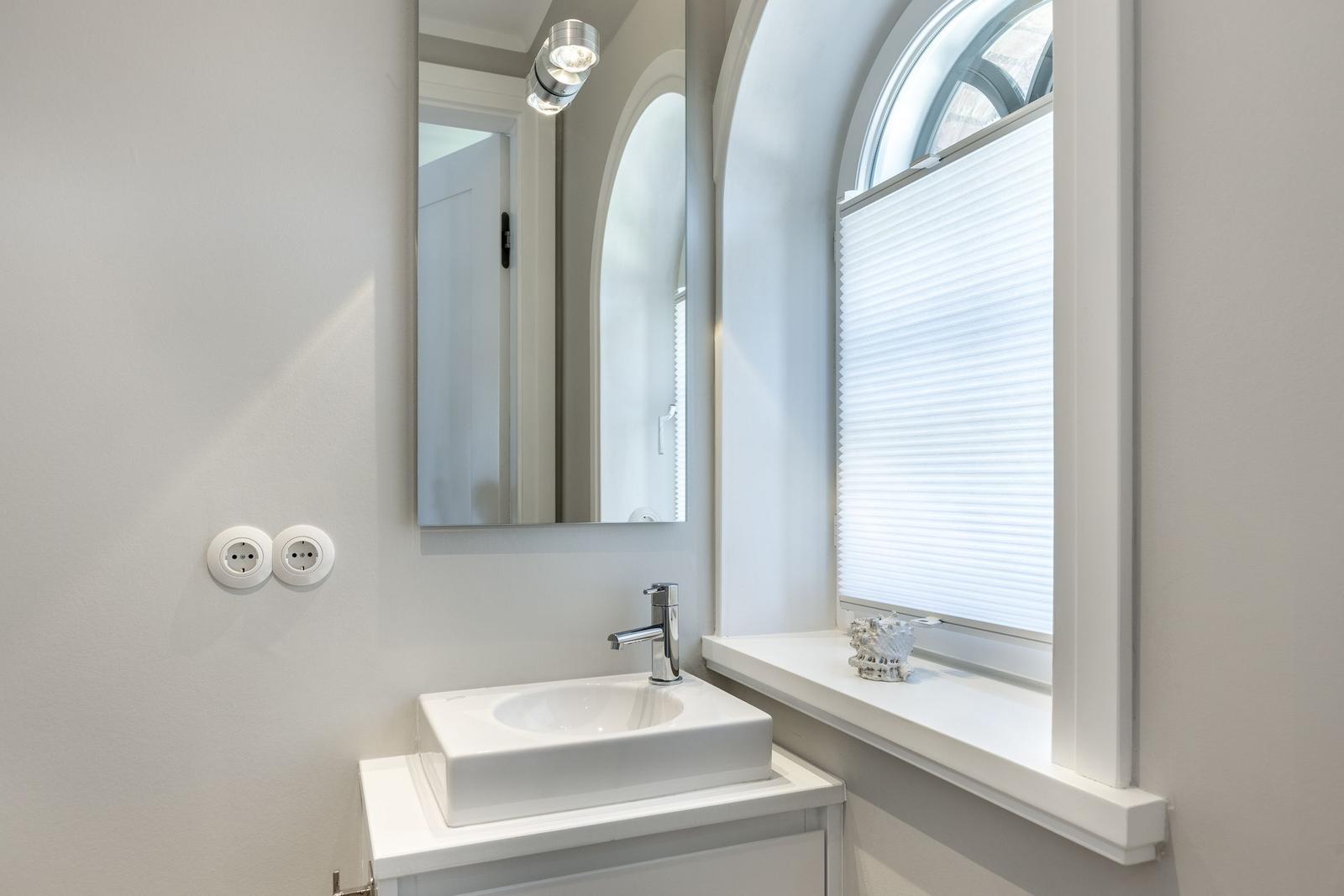 Badezimmer mit Waschbecken, Spiegel und Fenster mit Jalousie.