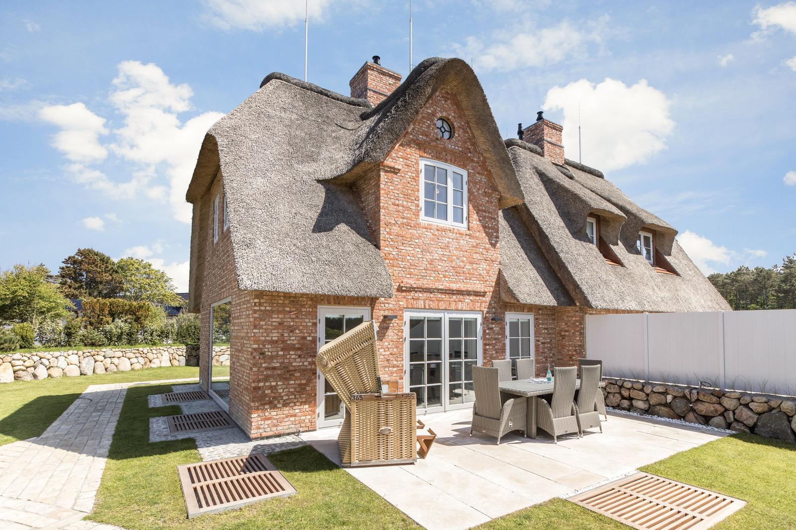 Haus mit Strohdach, Terrasse und Garten. Möblierte Außenterrasse mit Tisch und Stühlen.
