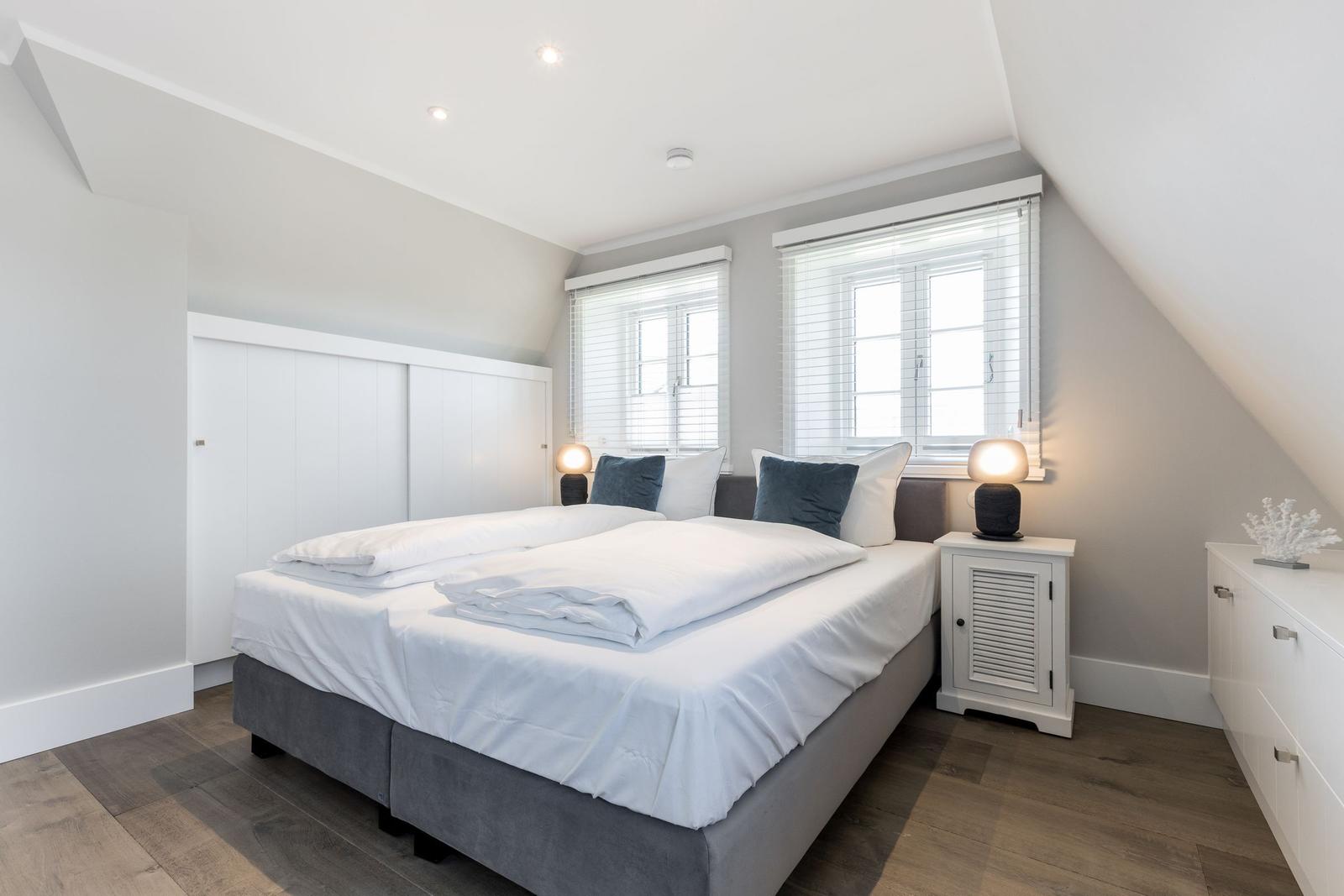 Schlafzimmer mit Doppelbett, zwei Fenstern und Nachttischen.