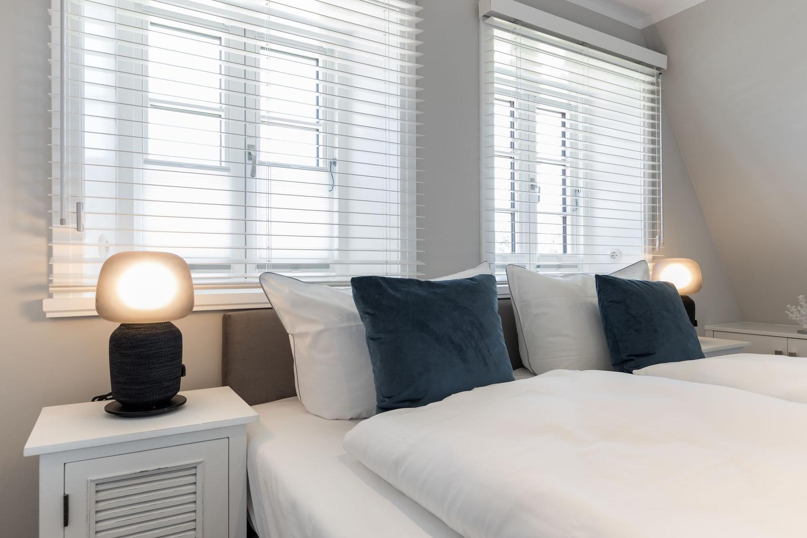 Schlafzimmer mit Doppelbett, zwei Nachttischen und Lampe. Fenster mit Jalousien.