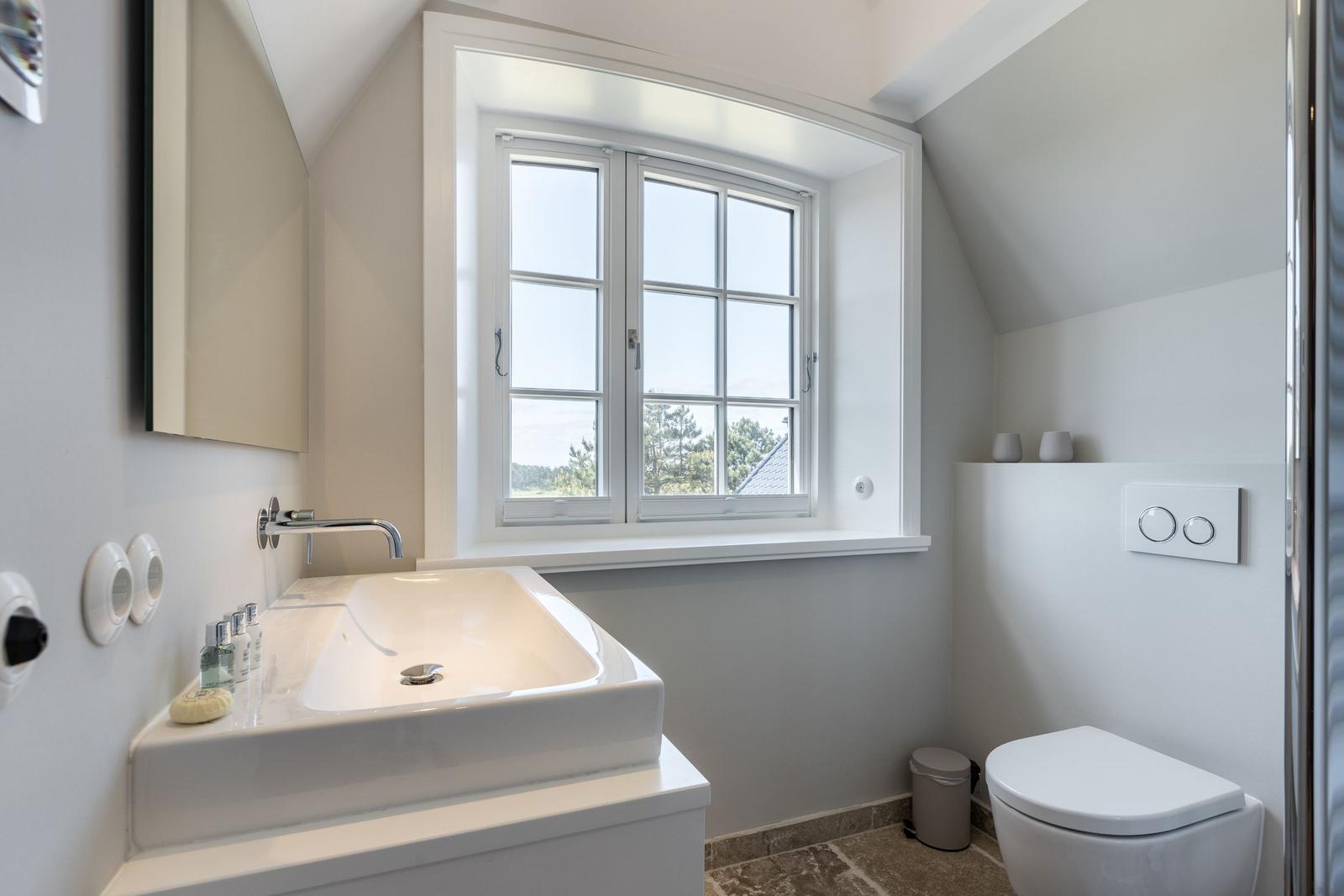 Badezimmer mit Waschbecken, Toilette und Fenster mit Blick auf Wald.