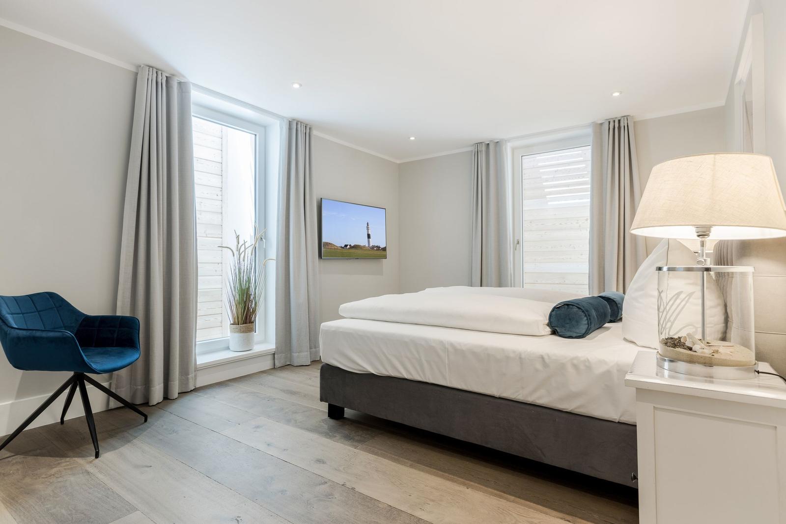 Schlafzimmer mit Bett, blauer Sessel und Fenstern.