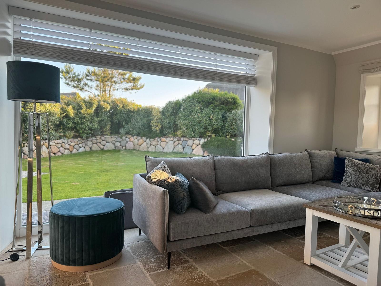 Großes Wohnzimmer mit grauem Sofa, blauem Hocker und Blick auf den Garten.