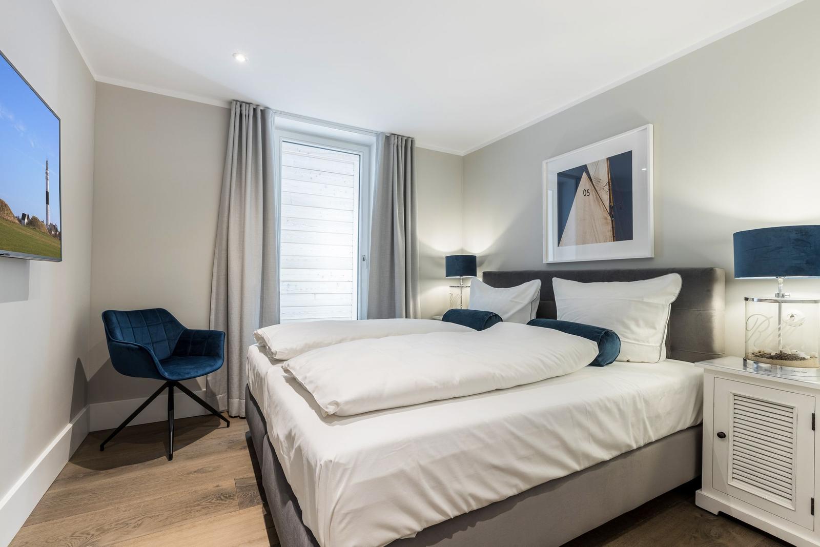 Schlafzimmer mit Doppelbett, blauer Sessel und Fenster mit Jalousie.