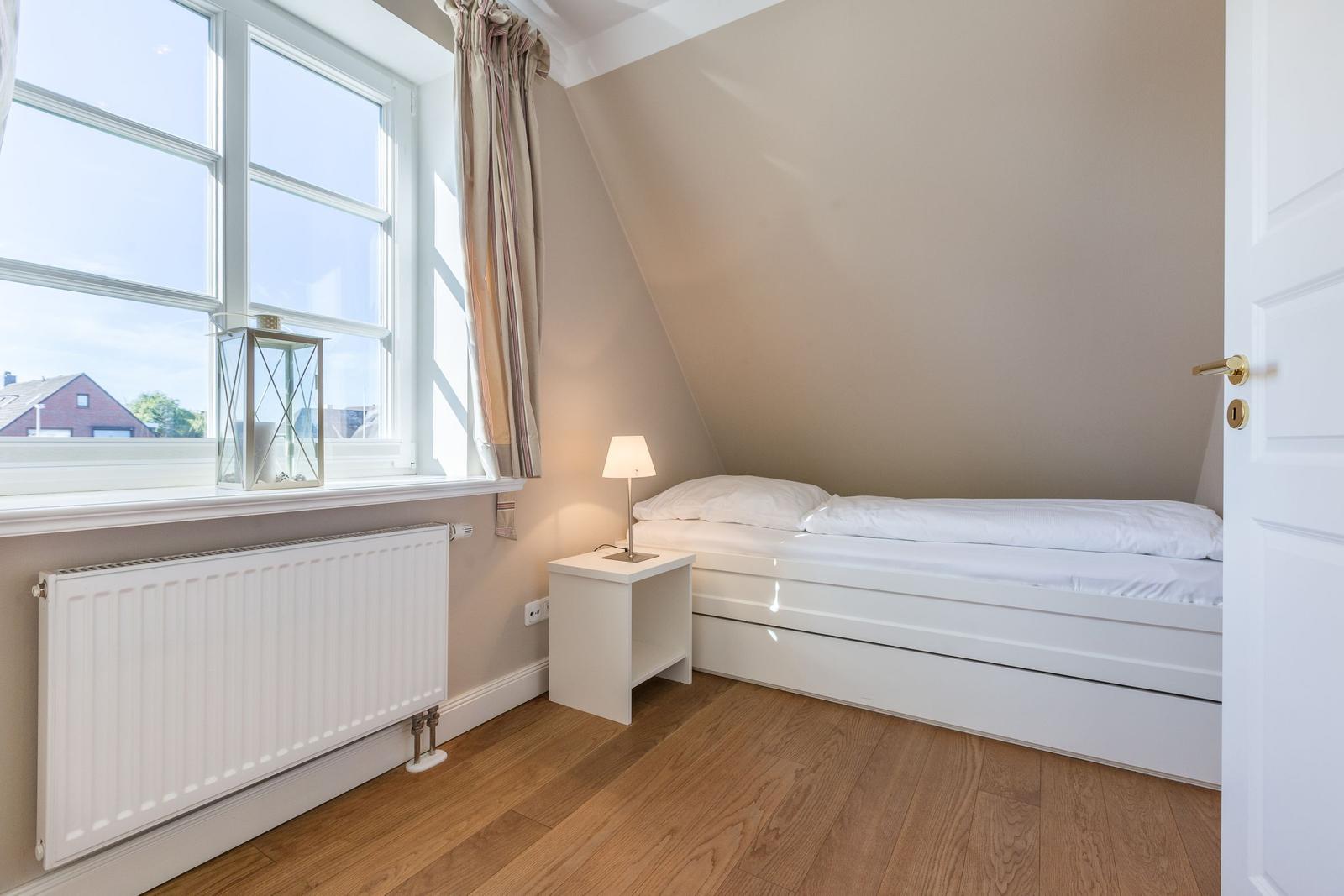 Schlafzimmer mit Dachschräge, weißem Bett und Holzboden. Fenster mit Vorhang und Heizkörper.