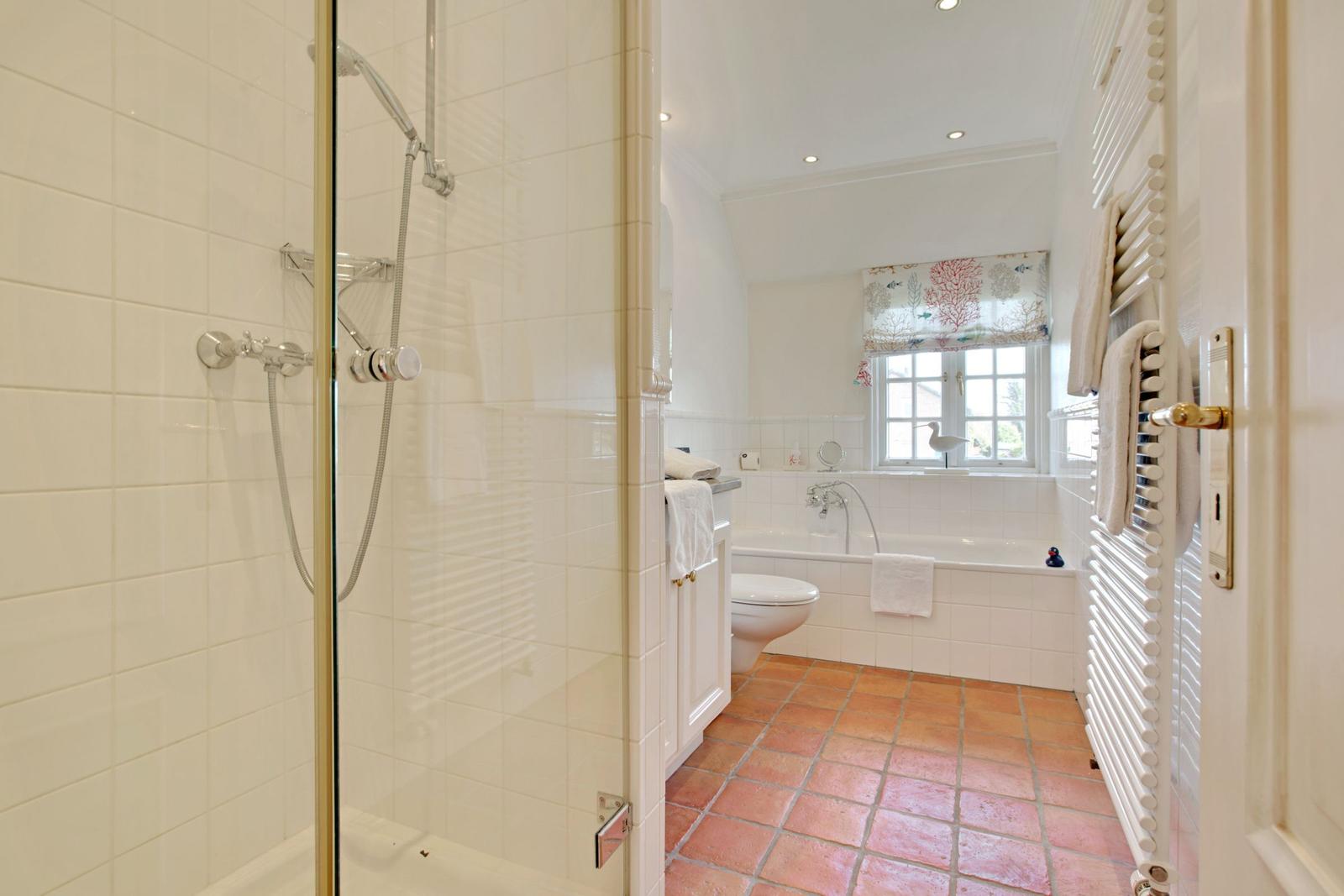 Badezimmer mit Dusche, Badewanne und Toilette. Terrakottafußboden und Fenster mit Vorhang.