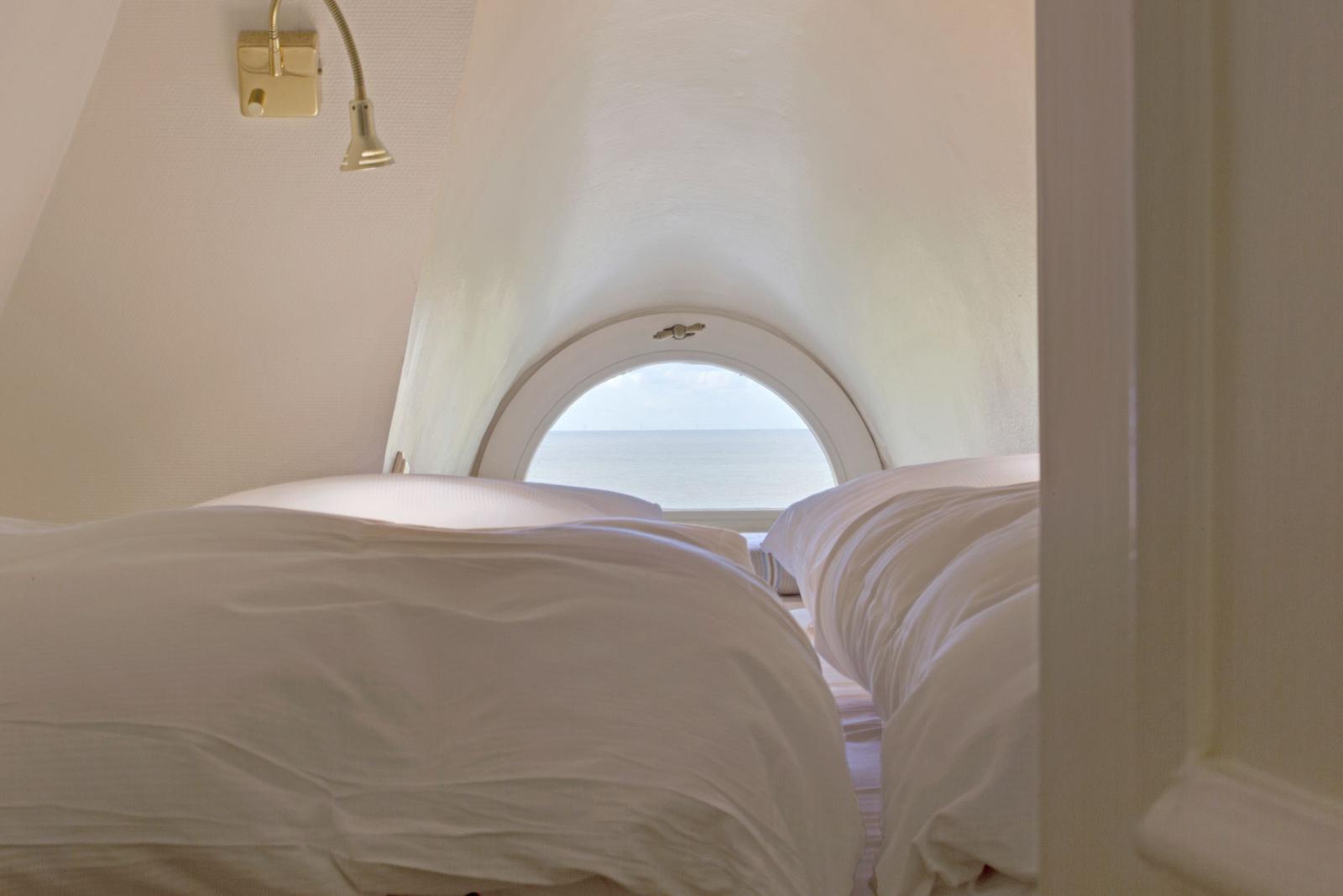 Zimmer mit Blick auf Meer durch runden Fenster, Bett mit weißen Bettwäsche, Wandlampe