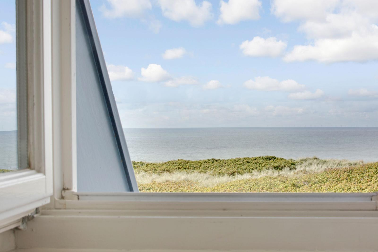 Durch das Fenster blickt man auf das Meer und grüne Dünen.