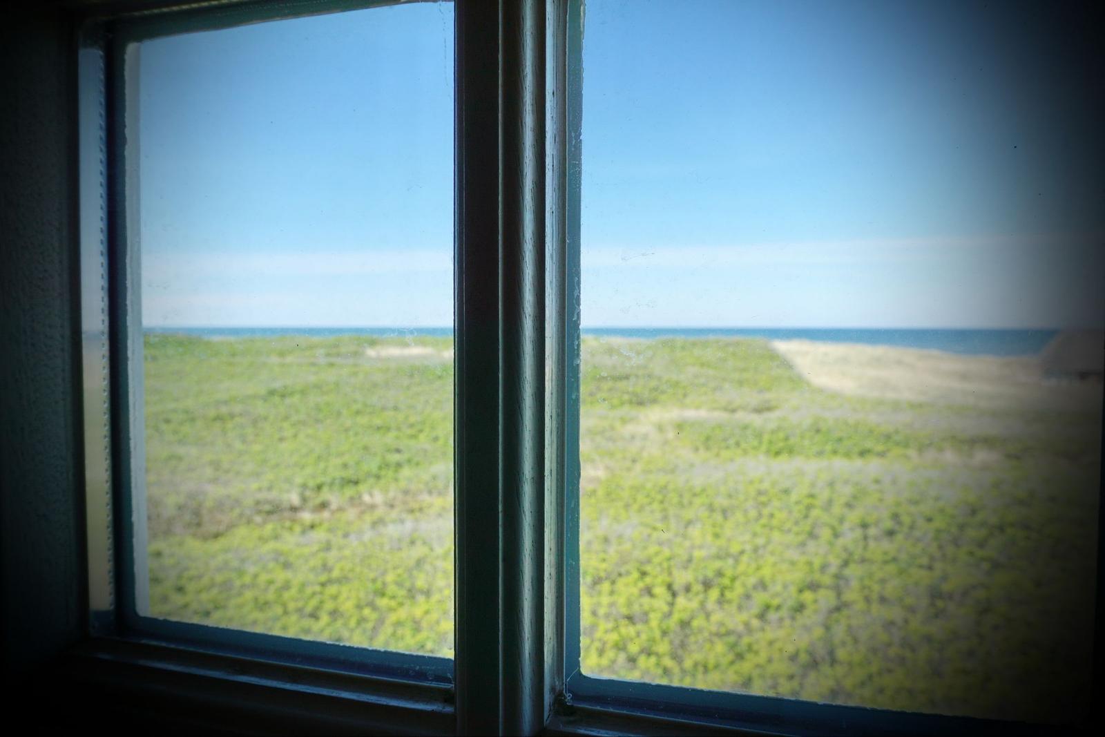 Durch das Fenster blickt man auf eine grüne Dünenlandschaft und das Meer.