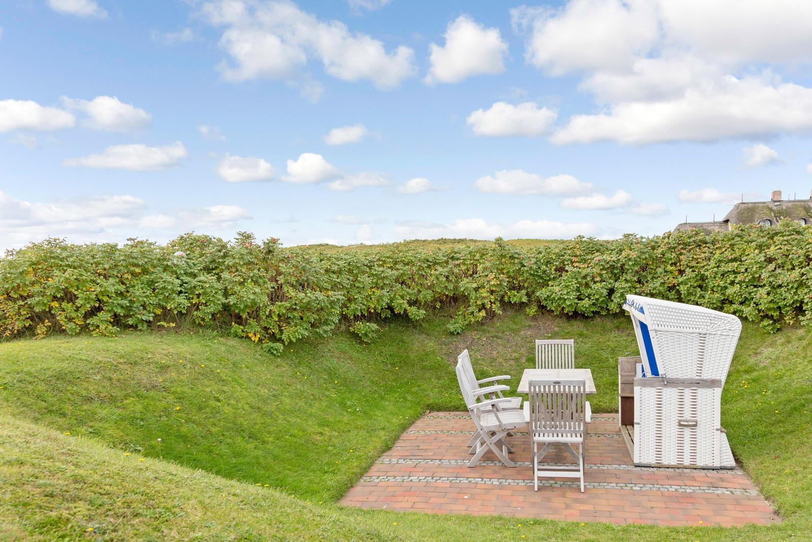 Terrasse mit Tisch, Stühlen und Strandkorb auf grünem Rasen unter blauem Himmel.