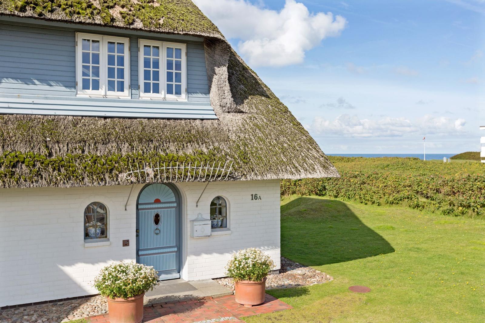 Haus mit Strohdach, weißer Fassade und blauer Tür. Grasfläche und Blick zum Meer.