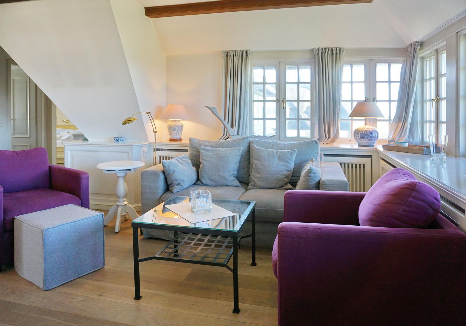 Wohnzimmer mit grauem Sofa, zwei violett gefärbten Sesseln und Glas-Tisch. Holzfußboden und große Fenster.