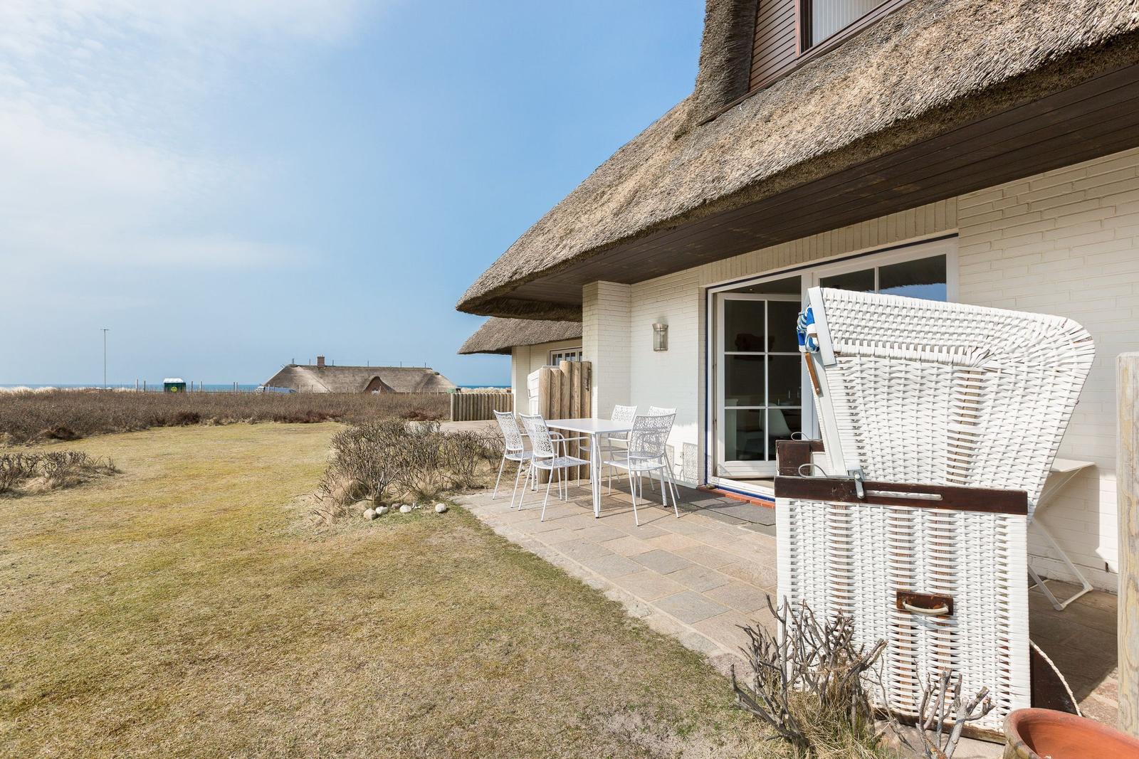 House with thatched roof, terrace with table and chairs, white wicker basket, view of beach and horizon.
