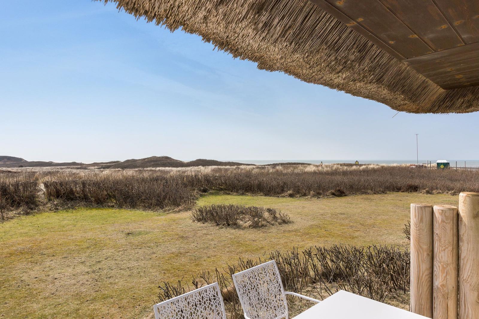 Terrace with thatched roof and white chairs overlooks dunes and beach.