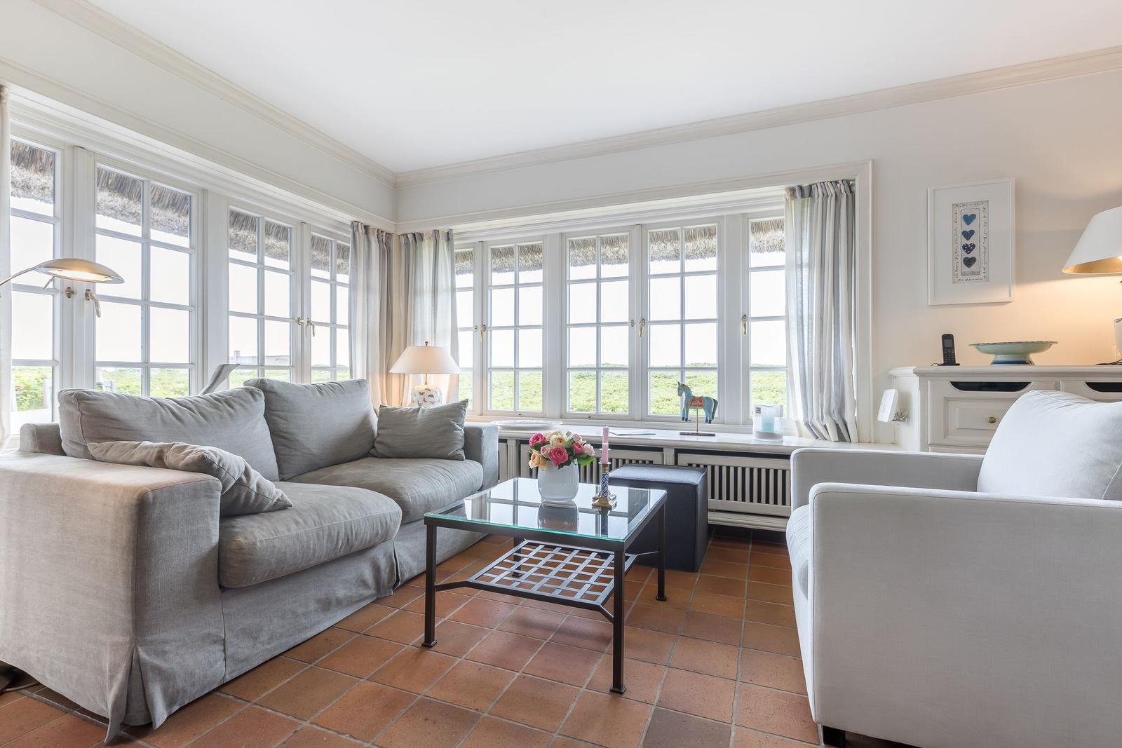 Wohnzimmer mit Sofa, Sessel und Glas-Tisch. Große Fenster mit Blick auf Grünfläche.