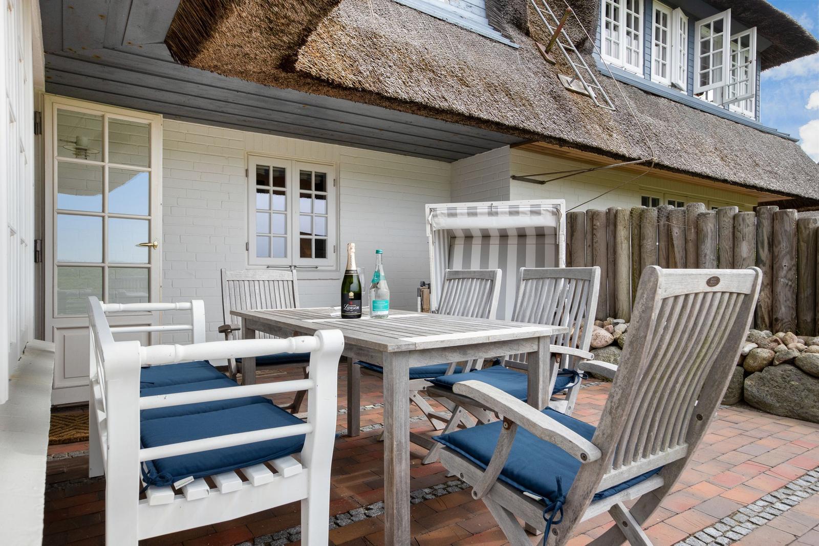 Terrasse mit Holztisch, Stühlen und blauen Kissen unter strohgedecktem Dach.
