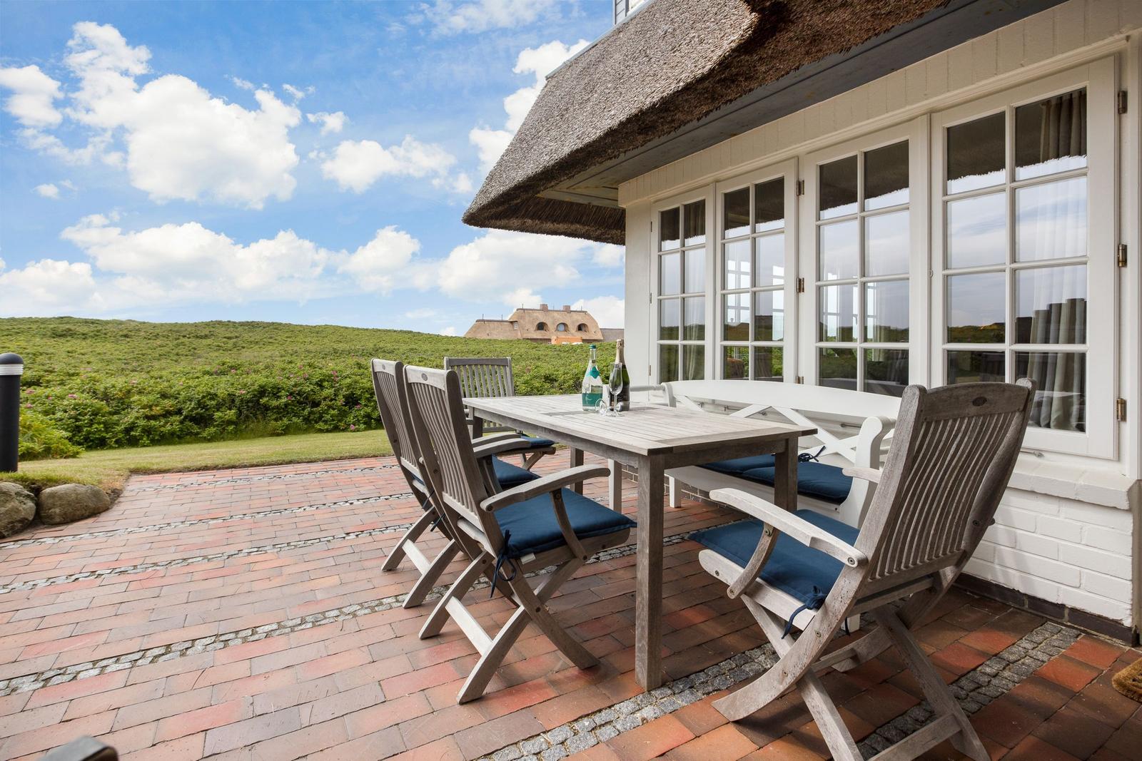 Terrasse mit Holztisch und Stühlen, Blick auf grüne Hügel und Himmel.