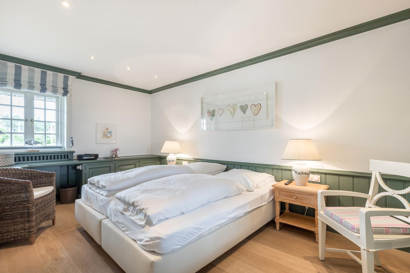 Schlafzimmer mit Doppelbett, Nachttisch und Stuhl. Fenster mit Vorhang. Wanddekoration.